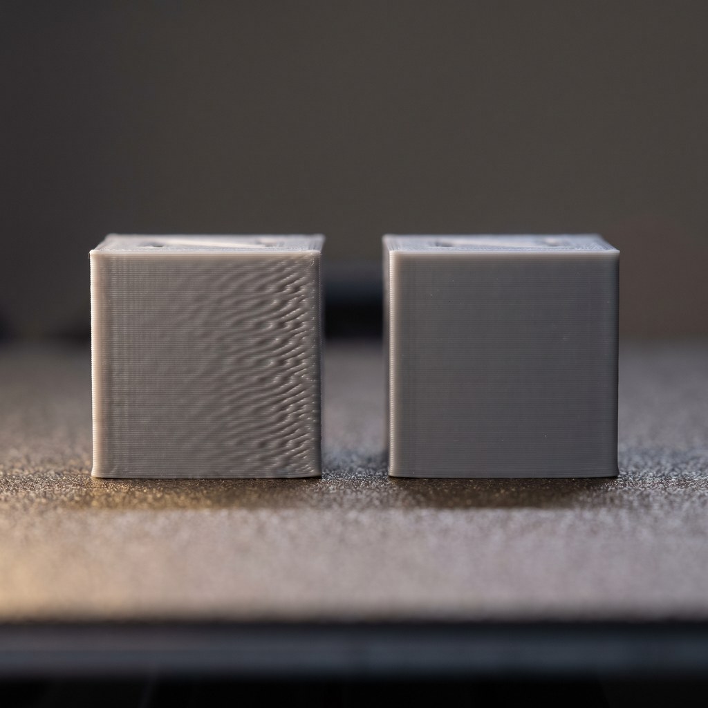 Dois cubos impressos em 3D lado a lado: o da esquerda com ondulações de ringing na parede, o da direita com superfície lisa